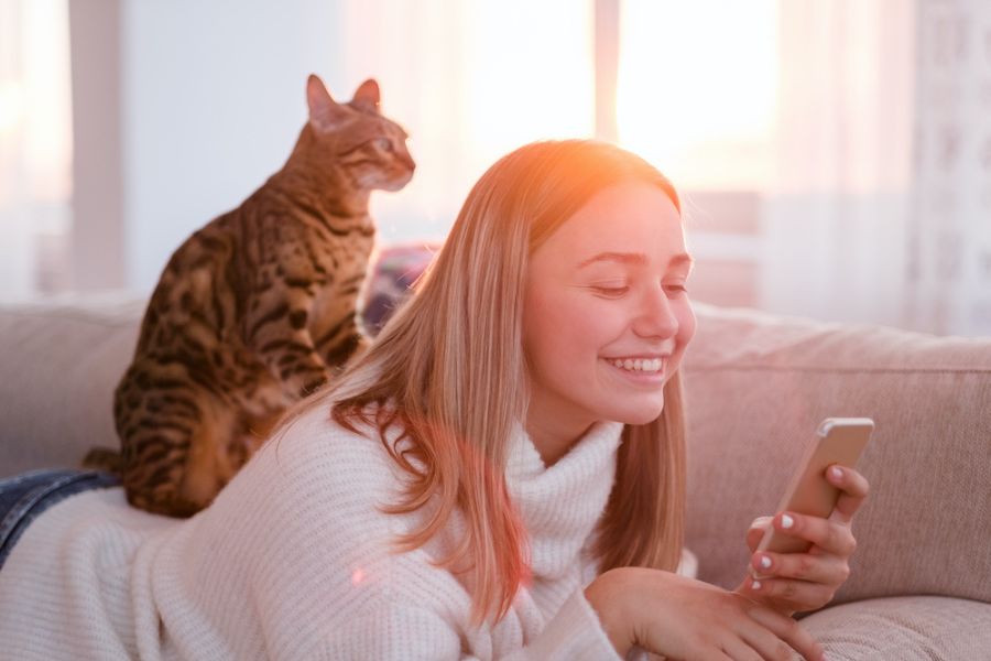 Un gatto appoggiato sulla schiena di una ragazza che sorride, con in mano uno smartphone