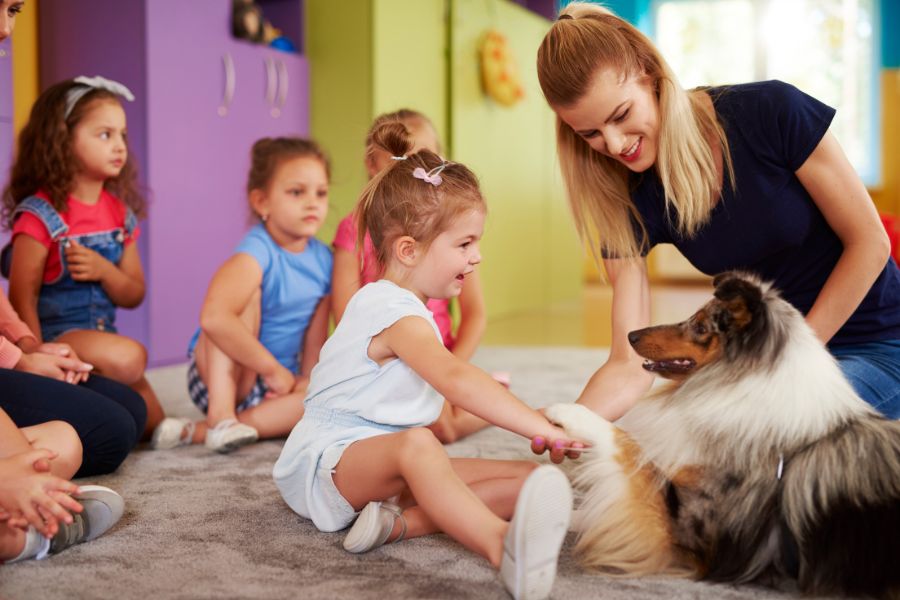 Una bambina gioca con un cane assieme a un'operatrice di pet therapy, sullo sfondo altri bambini